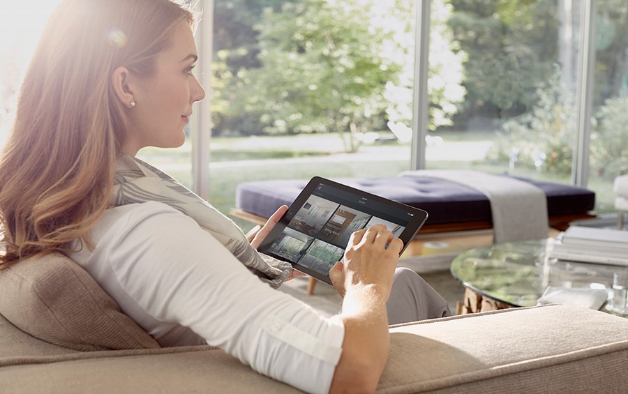 woman sits on couch while holding a tablet featuring a Savant home automation control interface